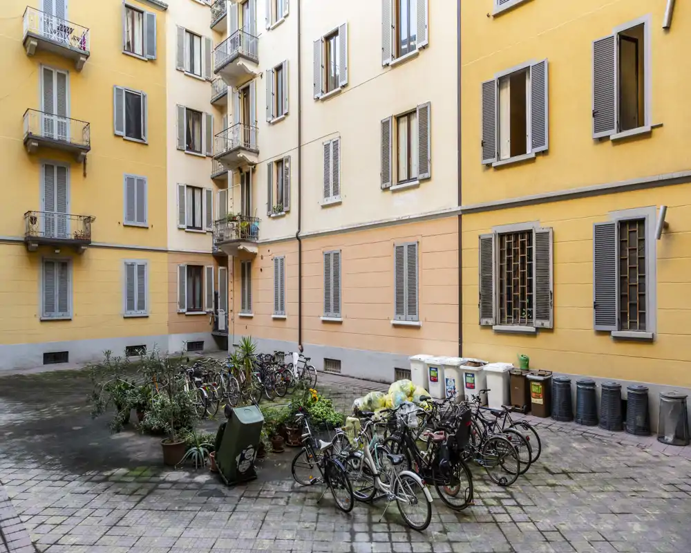 Cortile condominiale soleggiato con biciclette parcheggiate, vasi e cassonetti — ideale per annunci di appartamenti che mostrano gli spazi esterni condivisi.