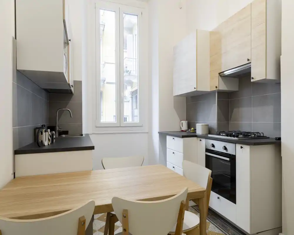 Cucina moderna e luminosa con tavolo in legno, quattro sedie, piano cottura a gas e forno incassato. Pensili neutri e paraschizzi piastrellato creano uno spazio pulito e funzionale, perfetto per affitti brevi.