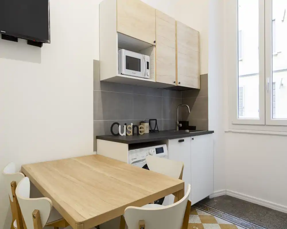 Cucina compatta e moderna con tavolo in legno, microonde incassato e lavello sul piano. Luce naturale intensa dalla finestra e finiture calde e pulite, perfetta per un piccolo appartamento.
