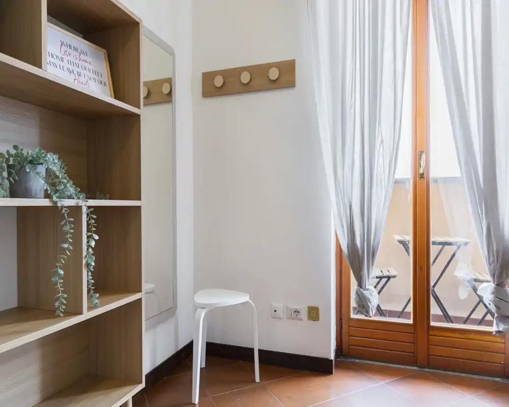 Ingresso luminoso e minimal con attaccapanni a parete, specchio a figura intera, scaffalatura e sgabello; porta a vetri che si apre su un piccolo balcone.