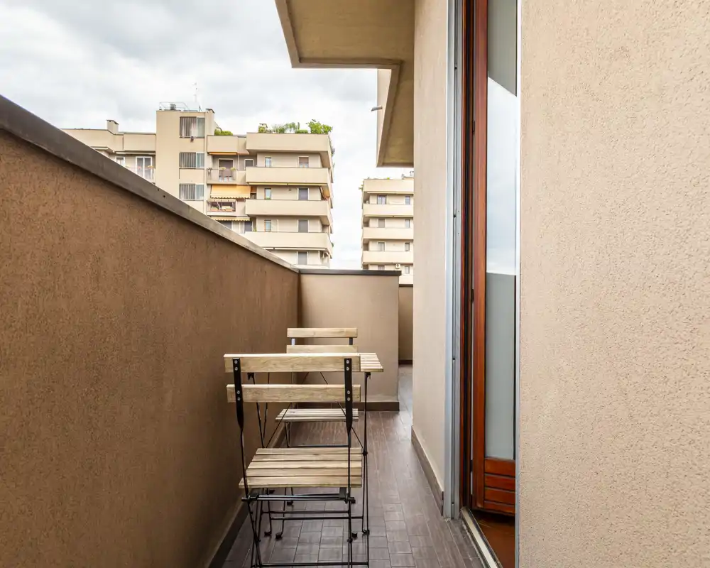 Balcone stretto con un piccolo tavolo bistro in legno e due sedie, pavimento piastrellato e vista sugli edifici vicini — ideale per il caffè mattutino.