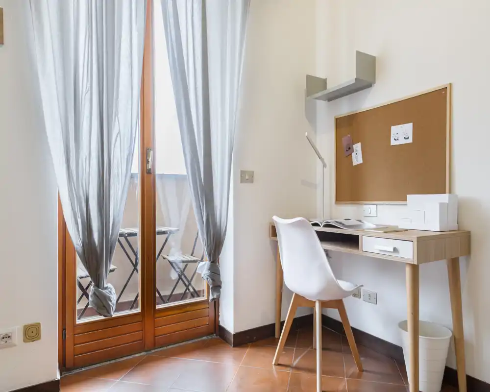 Angolo studio luminoso vicino alle porte-finestre con scrivania in legno chiaro, sedia moderna e bacheca in sughero; tende leggere e un piccolo balcone visibile.