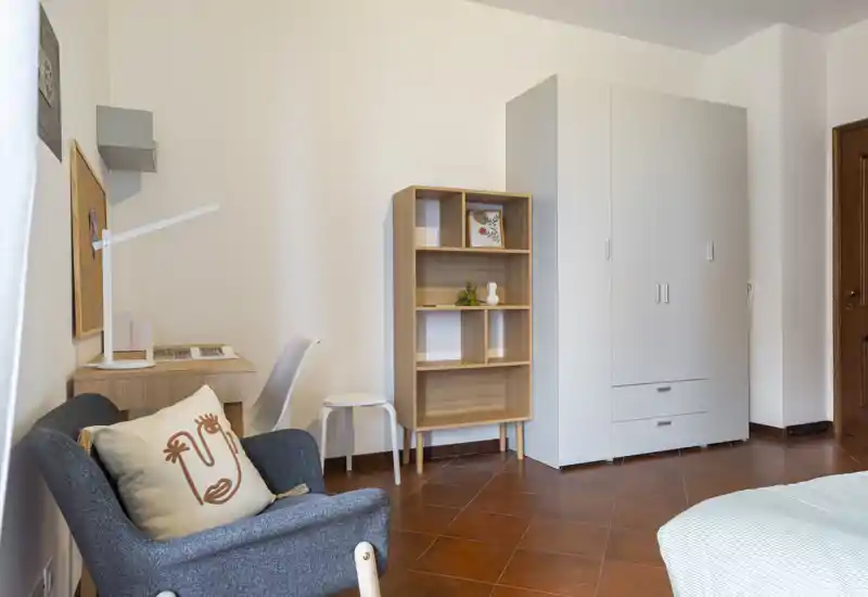 Angolo camera luminoso e ordinato con bordo del letto visibile, poltrona con cuscino, scrivania e armadio — arredo moderno e minimale perfetto per foto di affitto.