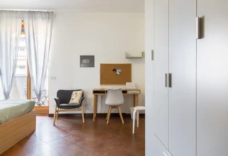 Camera da letto luminosa e ordinata con letto in legno, una scrivania con sedia e una poltrona accogliente vicino alla finestra; la luce naturale dal balcone illumina le piastrelle calde e l’arredamento minimal.