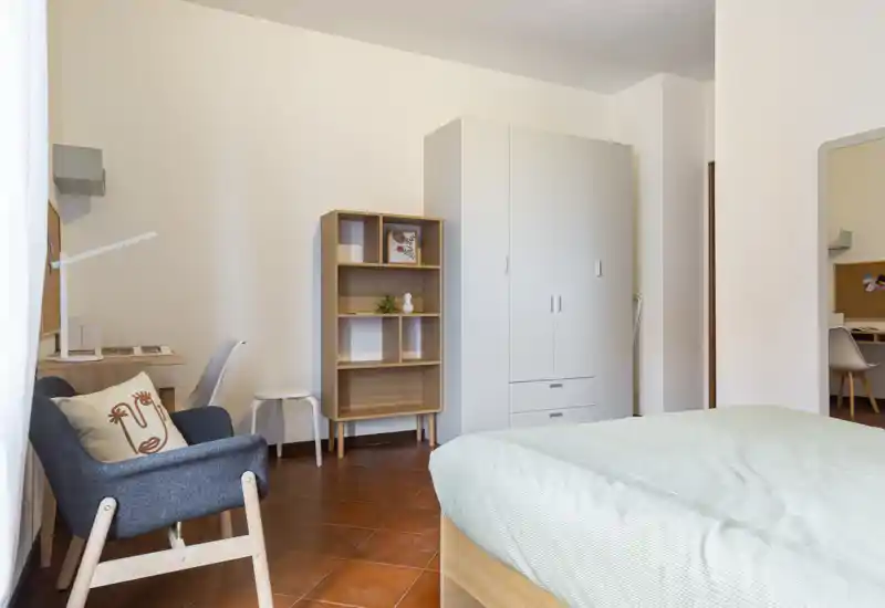 Camera da letto luminosa e ordinata con letto ben visibile, armadio, scaffale e una comoda poltrona. Luce naturale e pavimento caldo creano un'atmosfera accogliente, perfetta per gli annunci.