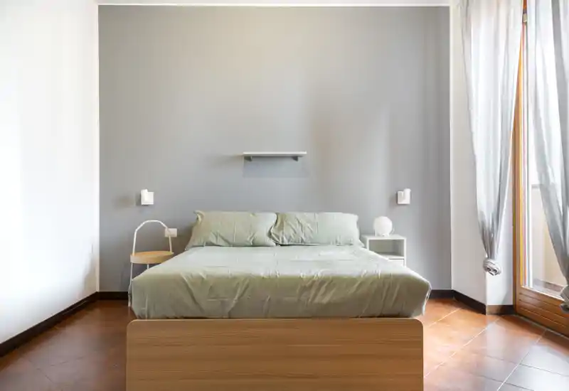 Camera da letto luminosa e minimalista con letto matrimoniale al centro, biancheria morbida e luce naturale da una grande finestra—spazio tranquillo e ordinato ideale per il riposo.