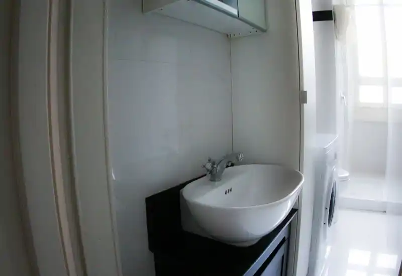 Salle de bain compacte et lumineuse avec lavabo posé moderne, robinetterie et machine à laver visible près de l'espace douche. Carrelage blanc et lumière naturelle depuis la fenêtre pour une atmosphère fraîche.