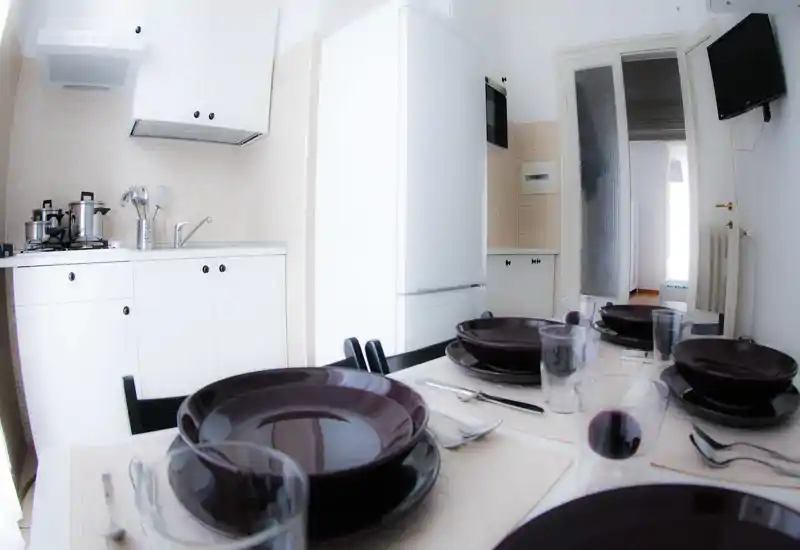 Cuisine compacte et lumineuse avec meubles blancs et réfrigérateur; la table est dressée avec assiettes foncées et verres, idéale pour présenter l'espace repas d'un petit appartement.