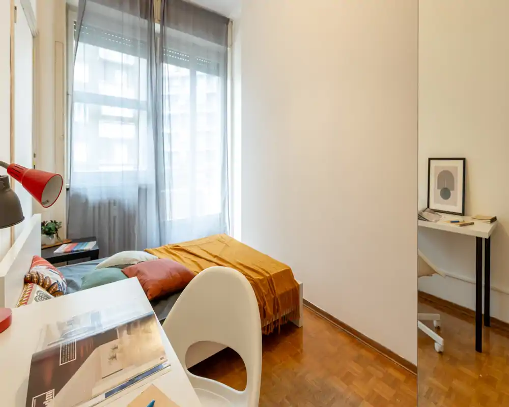 Habitación compacta y luminosa con cama individual, cojines y una cálida manta; un pequeño escritorio junto a la ventana grande proporciona luz natural y un espacio de trabajo práctico.