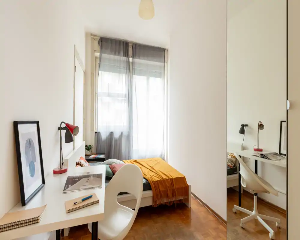 Dormitorio compacto y luminoso con cama individual, textiles cálidos, un escritorio sencillo y luz natural de una gran ventana — ideal para estudiante o persona sola.