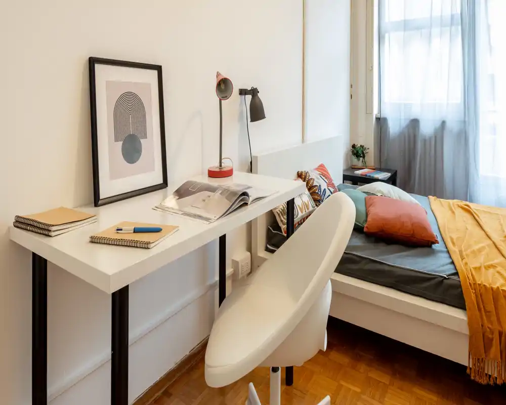 Dormitorio luminoso y con estilo con una cama individual con cojines de colores y una manta mostaza; un escritorio compacto y una silla moderna crean un espacio de trabajo práctico junto a la ventana.