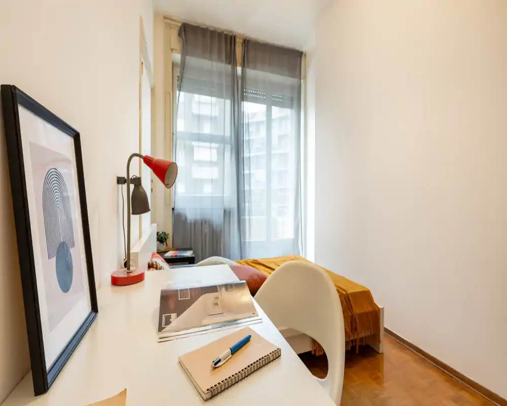 Dormitorio compacto y luminoso con cama individual, escritorio y ventana con cortinas translúcidas — ideal para estudiante o una sola persona.