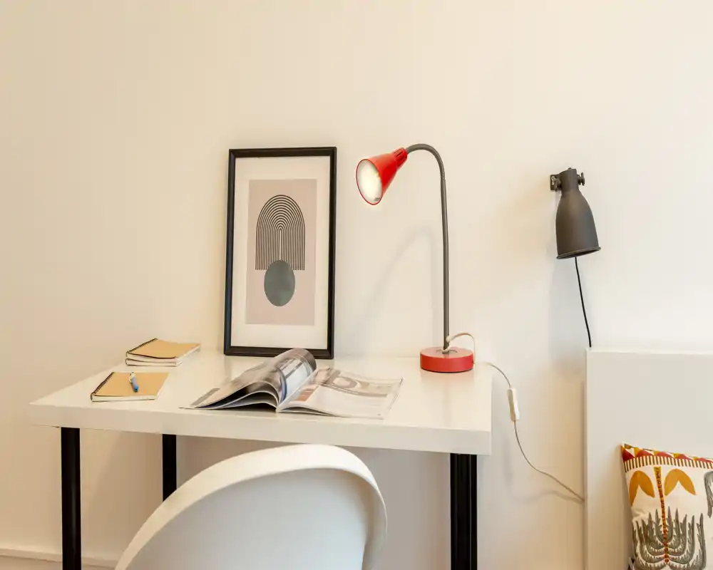 Rincón de dormitorio luminoso y minimalista con un escritorio blanco, una lámpara roja y un cuadro decorativo; se aprecia un pequeño fragmento de la cama/cabecero y un cojín a la derecha.