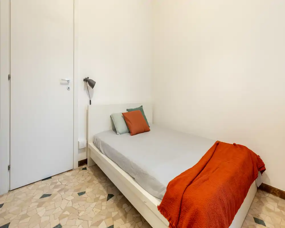 Dormitorio individual sencillo y luminoso con cama ordenada, cojines decorativos y una cálida manta naranja; paredes neutras y suelo de terrazo para un aspecto limpio y minimalista.