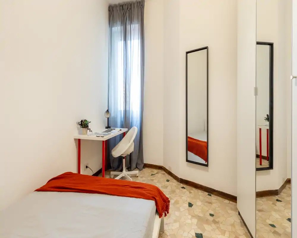 Dormitorio compacto y luminoso con cama individual con manta naranja, una pequeña zona de trabajo junto a la ventana y un espejo de cuerpo entero — limpio, minimalista y bien iluminado.