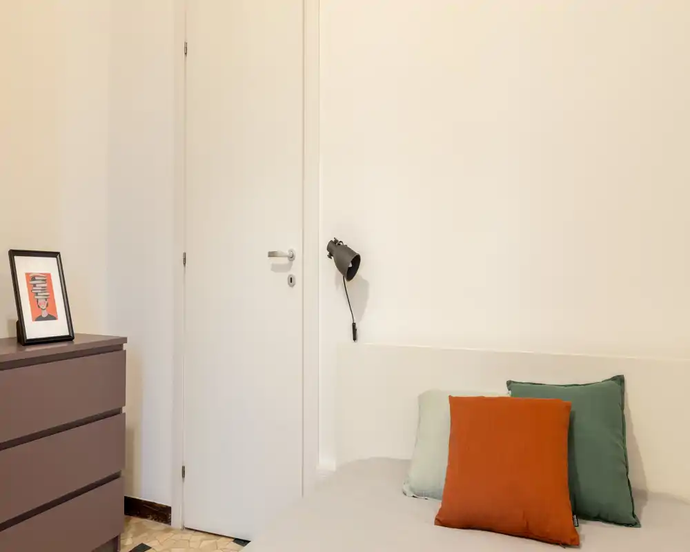 Rincón de dormitorio luminoso y minimalista con cabecero visible, cojines decorativos y lámpara de pared. Estilo limpio y moderno, perfecto para anuncios.