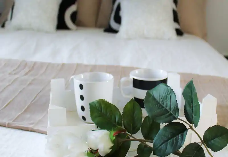 Camera da letto luminosa e curata con letto ben rifatto, cuscini decorativi e un vassoio con tazze e fiori — perfetta per un annuncio accogliente.