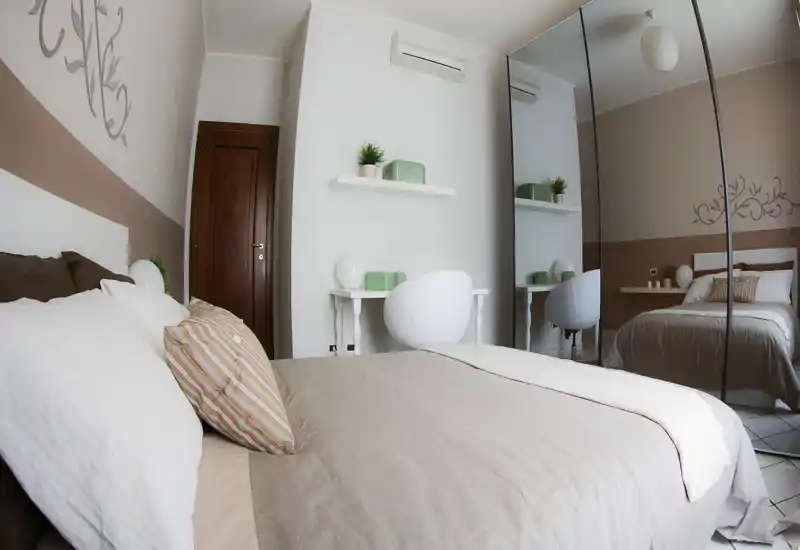 Camera da letto moderna e luminosa con un letto matrimoniale ben rifatto in primo piano, biancheria dai toni neutri e un armadio con ante a specchio che amplifica lo spazio.