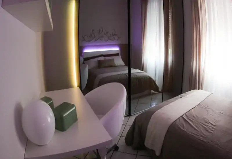 Camera da letto moderna e accogliente con letto matrimoniale, testiera decorativa e armadio con ante a specchio che riflettono la stanza. Luce naturale attraverso le tende leggere e illuminazione LED discreta per atmosfera.