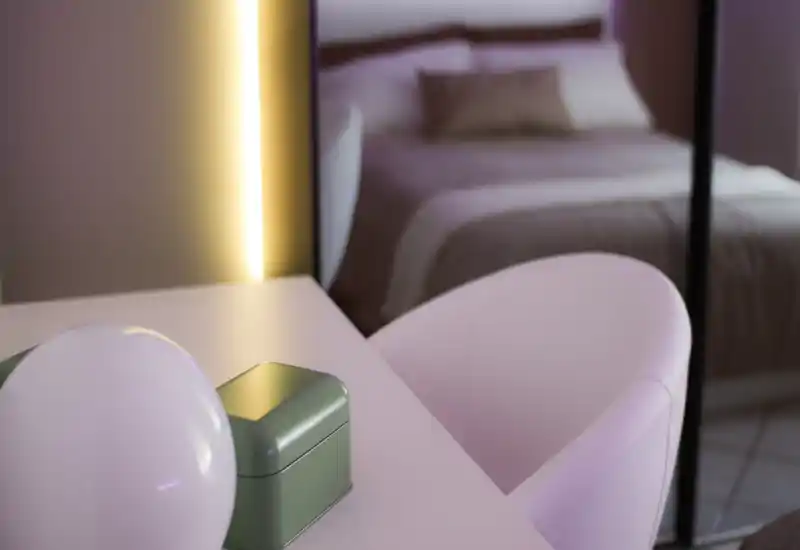 Scena di camera da letto in lieve sfocatura: il letto è visibile sullo sfondo, illuminazione calda verticale e arredi pastello in primo piano per un'atmosfera moderna e tranquilla.