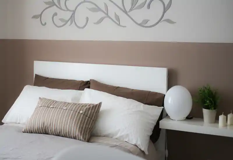 Camera da letto luminosa e curata con testiera bianca, cuscini sovrapposti e tonalità calde; una lampada da comodino e una piccola pianta rendono l'ambiente accogliente e moderno.