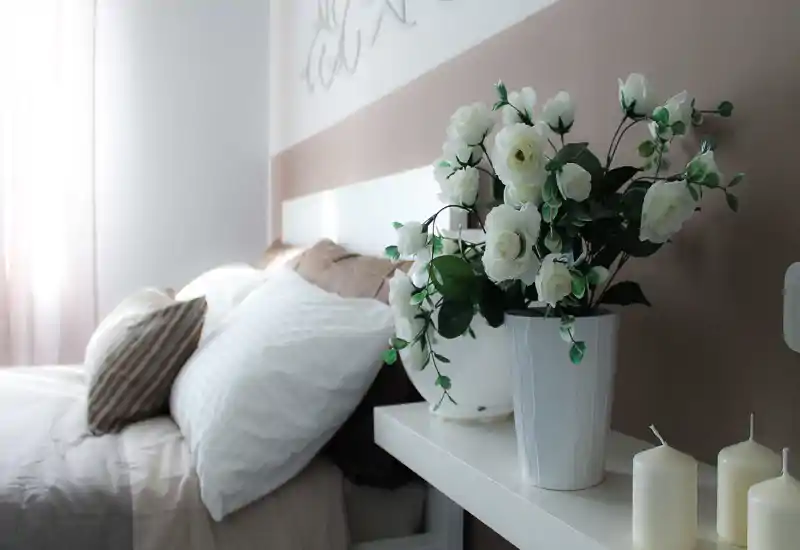 Camera da letto luminosa e dallo stile morbido, con testiera visibile e cuscini ordinati; fiori decorativi e candele sul ripiano del comodino donano un tocco accogliente ed elegante.