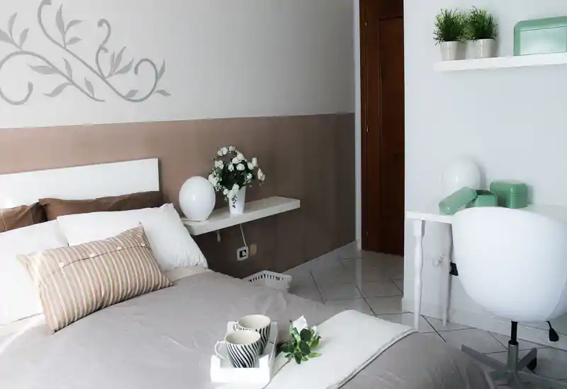 Camera da letto luminosa e curata con letto rifatto, cuscini decorativi e una piccola scrivania. Toni neutri e piante creano un'atmosfera accogliente, perfetta per annunci di affitto.