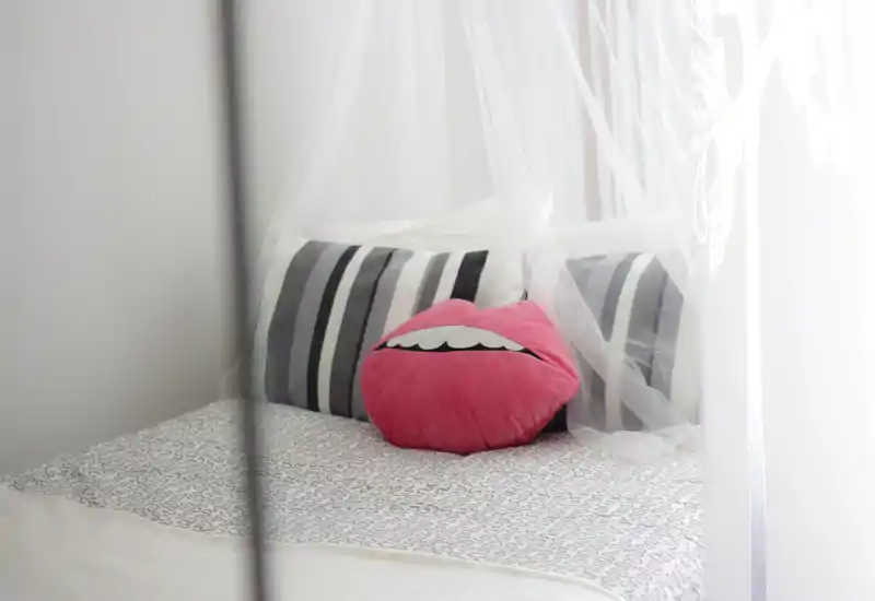 Rincón de dormitorio luminoso y de estilo suave con cama visible, cojines a rayas y un divertido cojín rosa de labios bajo un dosel de tul: limpio y acogedor.