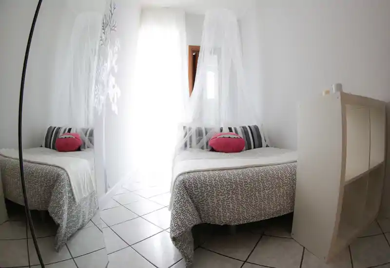 Dormitorio luminoso y aireado con cama doble, dosel decorativo y cojines a rayas; suelo de baldosa y armario con espejo que refleja la estancia.