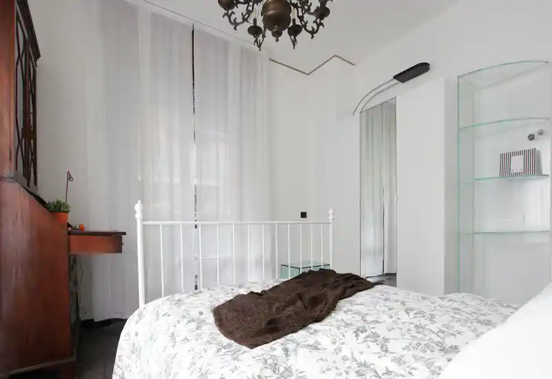 Dormitorio luminoso y bien compuesto con cabecero metálico blanco, ropa de cama con estampado y una manta acogedora. Las cortinas traslúcidas difuminan la luz natural y los muebles de madera aportan calidez.
