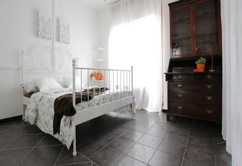 Dormitorio luminoso y aireado con cama metálica blanca, ropa de cama floral y una cómoda de madera oscura. Gran ventana con cortinas translúcidas que inunda la habitación de luz natural, creando un ambiente limpio y acogedor.