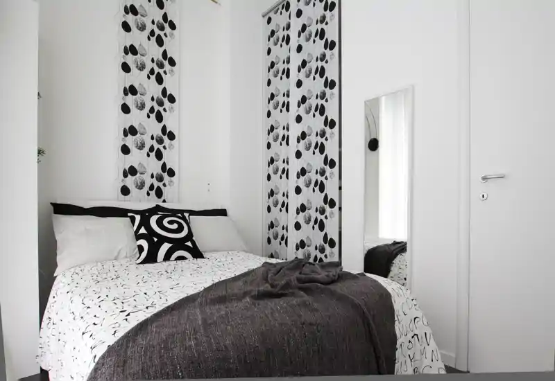 Bright, minimalist bedroom with a double bed dressed in patterned linens, decorative pillows and a dark throw; white walls and a full-length mirror create a clean, airy feel.
