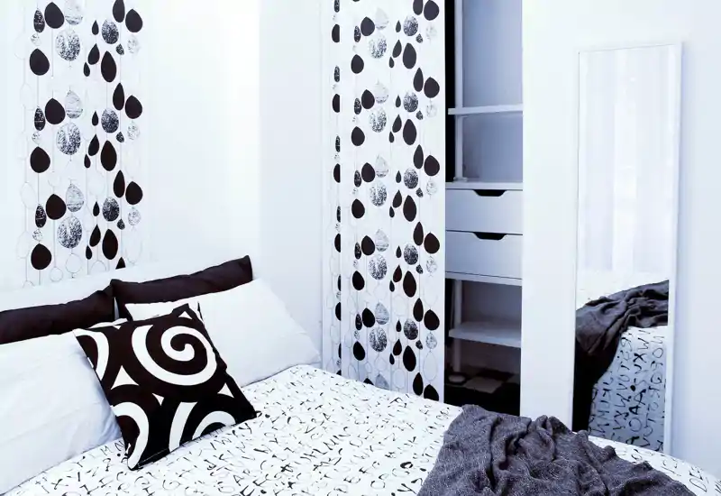 Dormitorio moderno y luminoso con textiles estampados, cojín decorativo y cama bien visible en primer plano. Diseño limpio en blanco y negro con espejo y armario empotrado.
