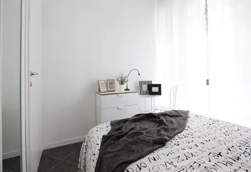 Bright, minimalist bedroom with a large bed in the foreground, white dresser, decorative frames and a cozy dark throw over patterned bedding. Natural light filters through sheer curtains creating an airy, inviting atmosphere ideal for listings.