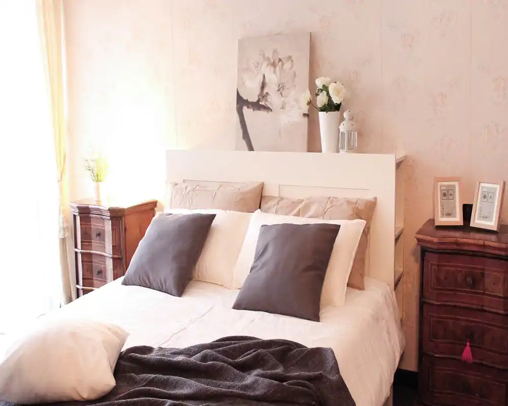 Bright, well-staged bedroom with a large double bed, neutral linens and decorative cushions. Soft natural light, vintage bedside cabinets and floral accents create a cozy, inviting atmosphere ideal for a rental listing.