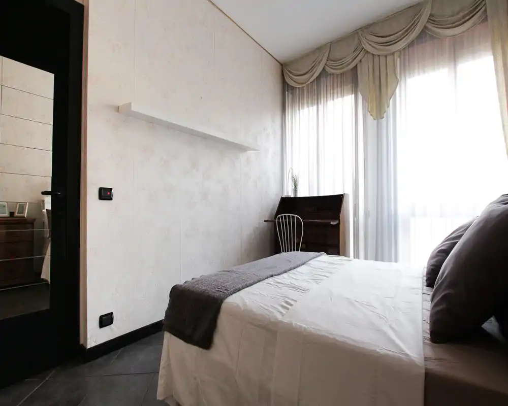 Bright, neatly staged bedroom with a double bed, soft linens and natural light from a large window. A small writing desk and chair sit by the window, creating a cosy corner.