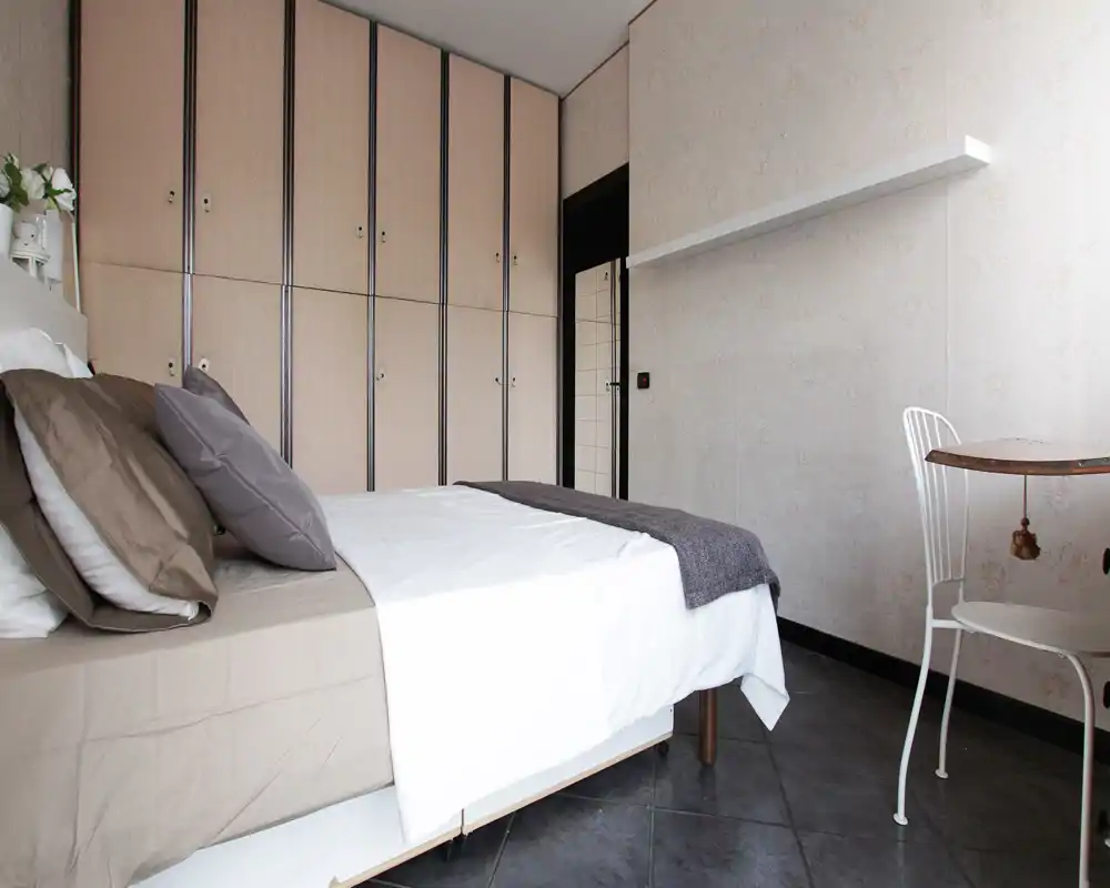 Bright, neatly made bedroom with a double bed, neutral linens and built-in wardrobe along the wall. The space is styled simply with coordinated pillows and a small table and chair, ideal for a restful stay.