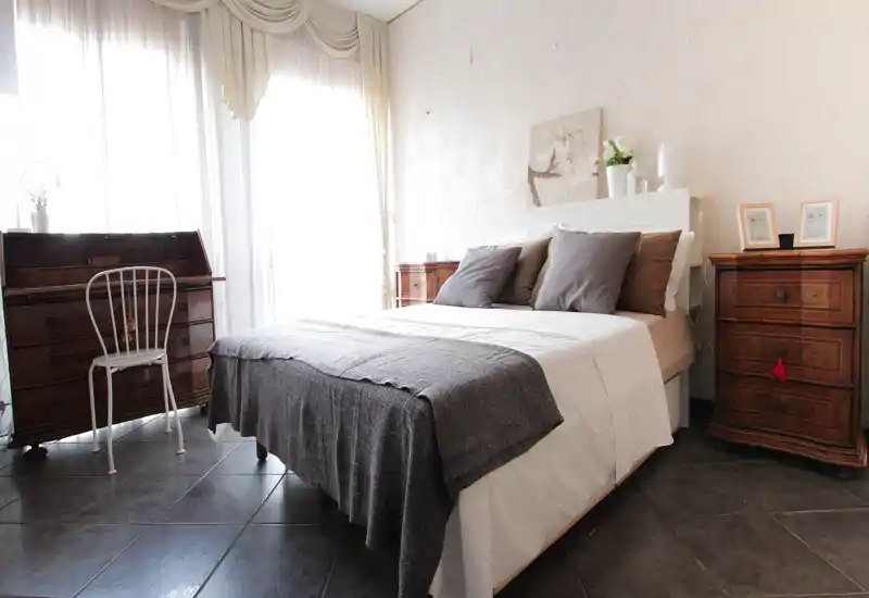 Bright, neatly styled bedroom with a centrally placed double bed, layered pillows and a soft throw, flanked by matching wooden bedside chests and a writing desk by the window.