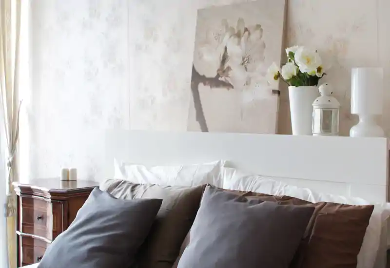 Bright, stylish bedroom with a visible white headboard, layered pillows and soft neutral decor; natural light from the window and decorative accents create a cozy, inviting atmosphere.