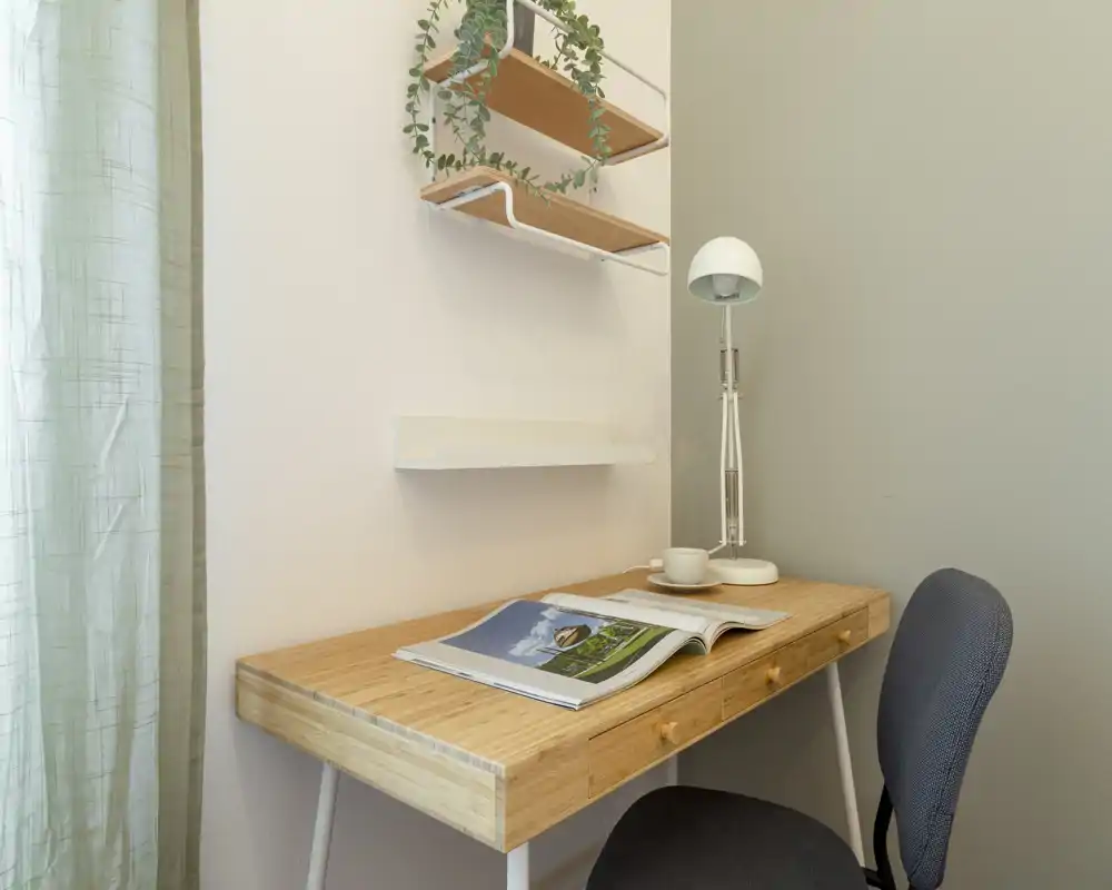 Compact, light-filled workspace with a wooden desk, reading lamp and a comfortable chair. Simple wall shelves and a small plant add a cozy touch.