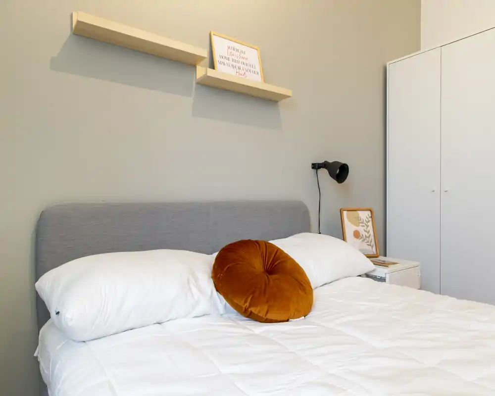 Bright, modern bedroom with a grey upholstered headboard, white bedding and a warm orange round cushion; minimalist shelving and a white wardrobe complete the scene.