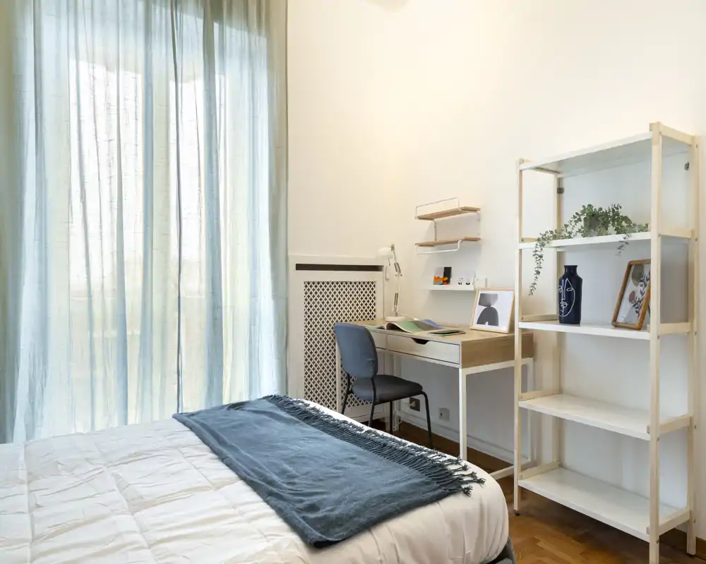 Bright, minimal bedroom with a neatly made bed, a study desk and open shelving—soft natural light filters through sheer curtains creating a calm, airy atmosphere.