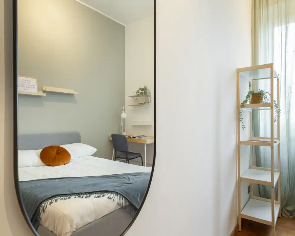 Bright, modern bedroom with a neatly made double bed seen through an elegant wall mirror; soft textiles, a round decorative cushion and a small desk create a cozy, functional space.