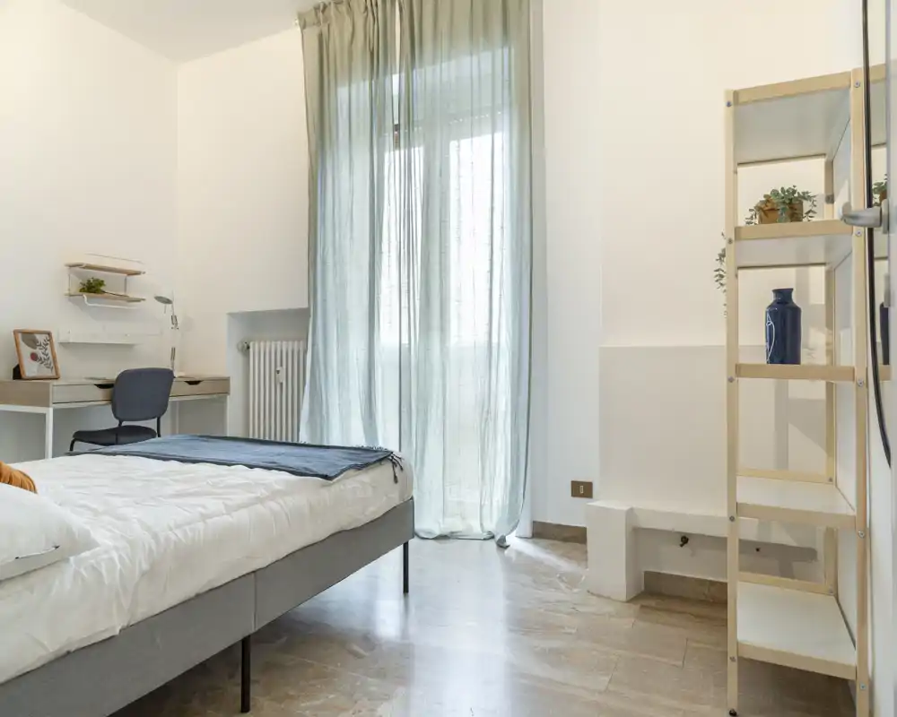 Bright, softly furnished bedroom with a double bed, sheer curtains and a small desk — calm minimalist styling and natural light.