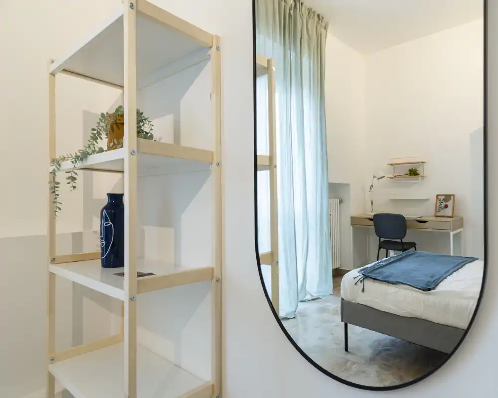 Bright, modern bedroom shown through a large wall mirror: a neatly made bed with a blue throw, a small desk and chair by the window, and soft natural light through sheer curtains.