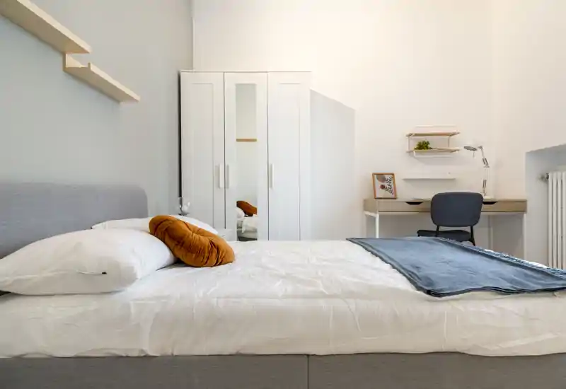 Bright, modern bedroom with a neatly made double bed, warm accent cushion and a small desk by the wall — ideal for a comfortable stay or remote work.