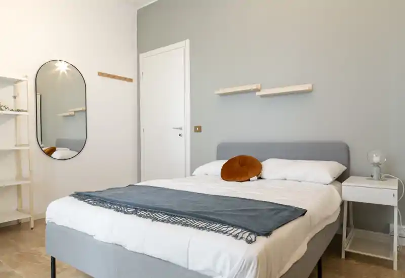 Bright, modern bedroom with a fully visible double bed dressed in white linens, a decorative rust-coloured round cushion and a blue throw. Minimal Scandinavian decor with a bedside table, oval wall mirror and soft grey accent wall create a calm, inviting atmosphere.