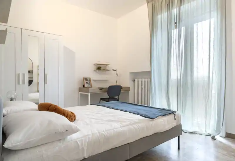 Bright, modern bedroom with a comfortable double bed, soft linens and a small desk by the window. Sheer curtains let in natural light and the neutral palette creates a peaceful atmosphere.