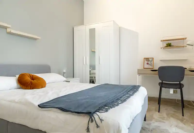 Camera da letto luminosa e ordinata con letto matrimoniale con biancheria bianca, plaid blu e cuscino arancione decorativo; armadio e scrivania compatti per una disposizione funzionale.