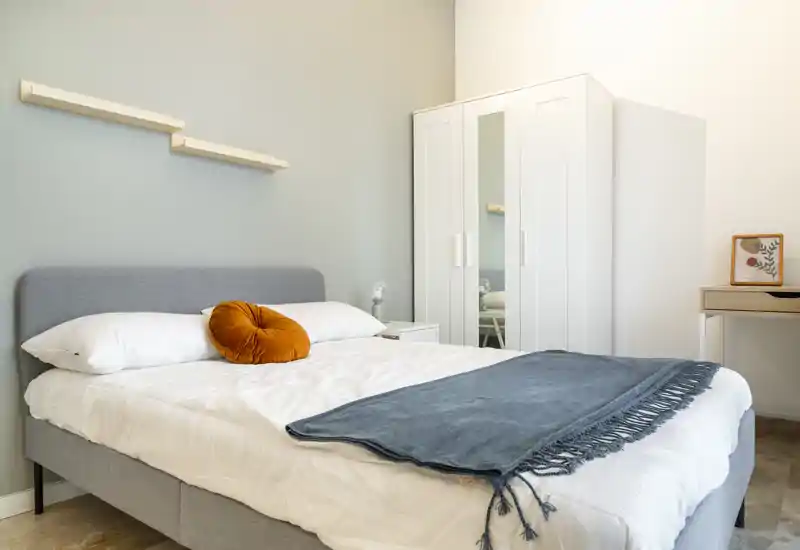 Sunny, modern bedroom with a neatly made double bed, soft gray headboard, decorative orange cushion and a blue throw. White wardrobe with mirrored door and a small desk complete the bright, minimal space.